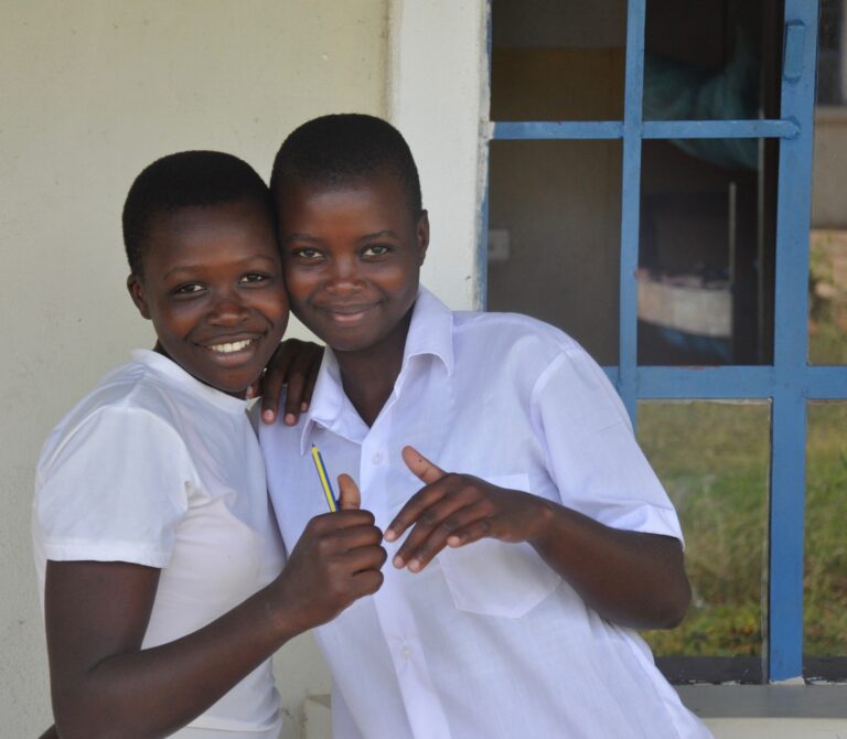 Two WISER girls hugging and posing for a picture. One girl is holding a pencil in her hand, and the other girl is holding her hand out.