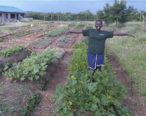 A girl is standing in a field with various crops growing. She has a big smile on her face and is holding out her arms.